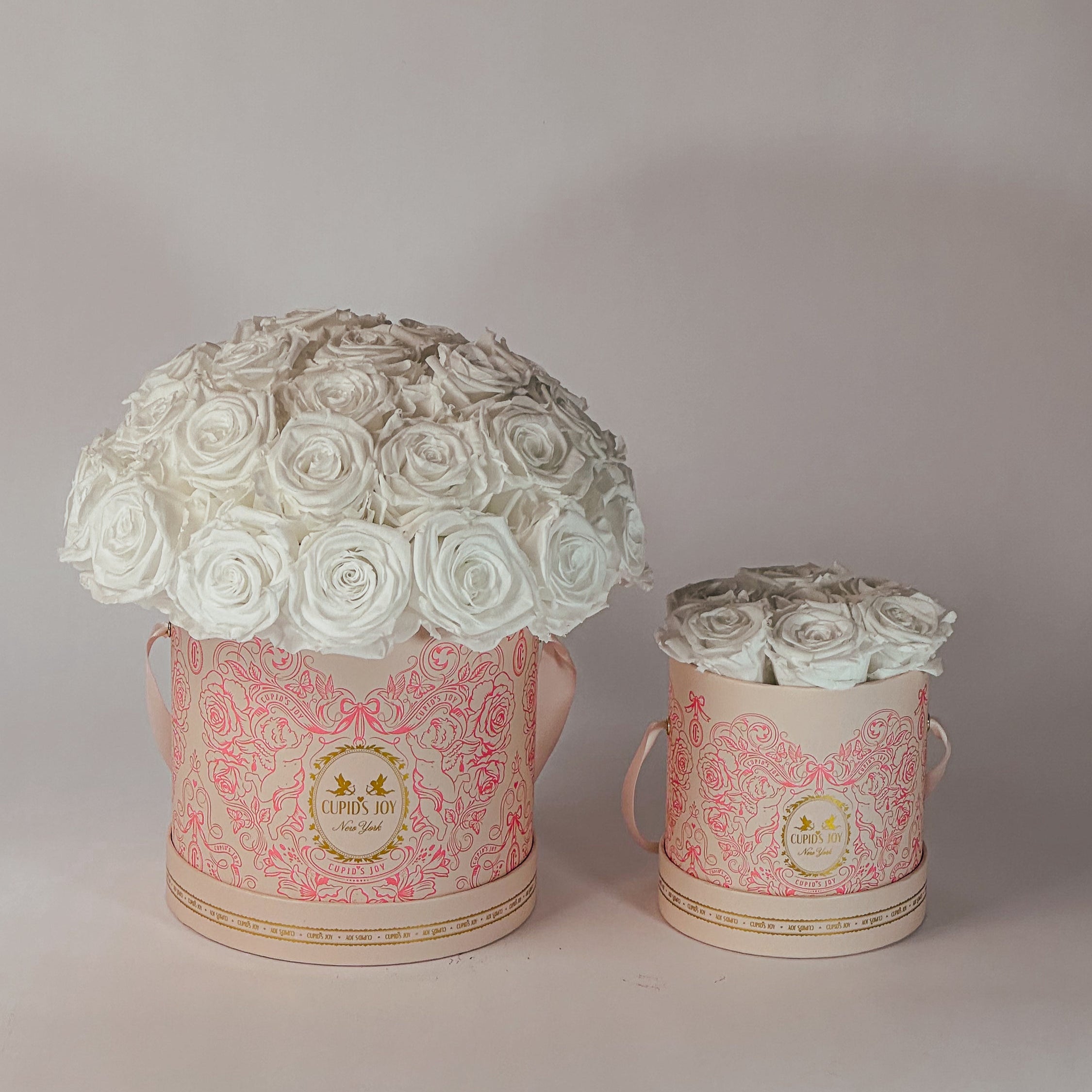 Two cupid-motif boxes by Cupid's Joy with white rose arrangements on a plain background